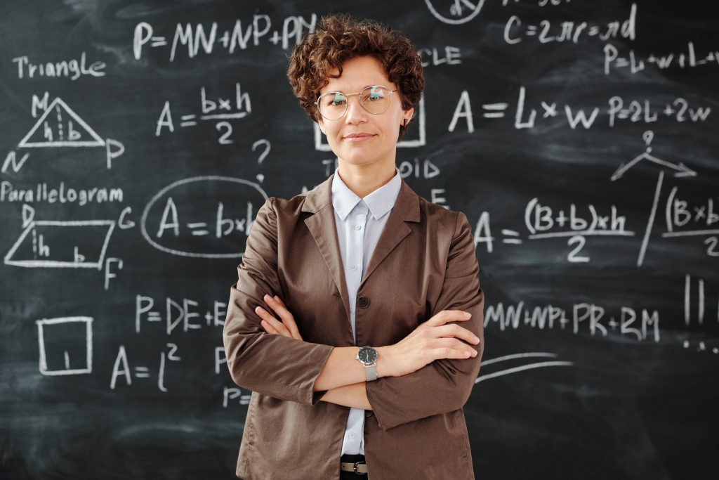 A young women dressed in a white t-shirt and a blazer, stands with crossed arms in front of a blackboard full of mathematical formulas. She looks steadily to the camera. This picture is used to represent the unequal mid-career trajectories which are studied in work package five