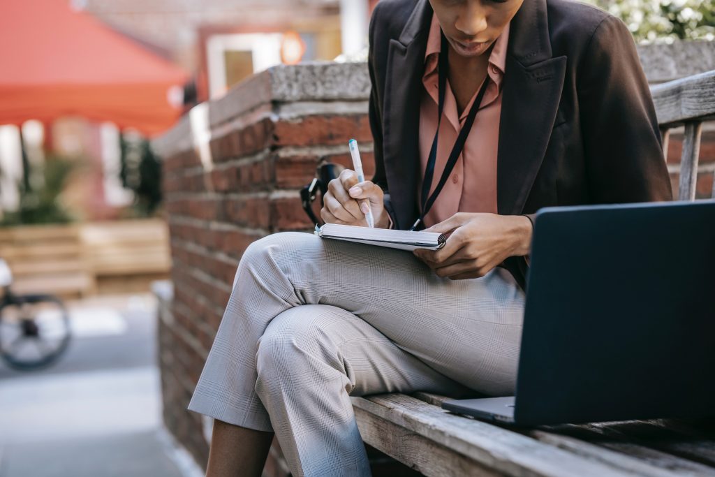 A women sits on a bench in the walks side. She takes notes in a notebook while looking at her laptop computer. The photo is used to illustrate the search for a job and the inequalities in the school-to-work transition which are studied in work package four.
