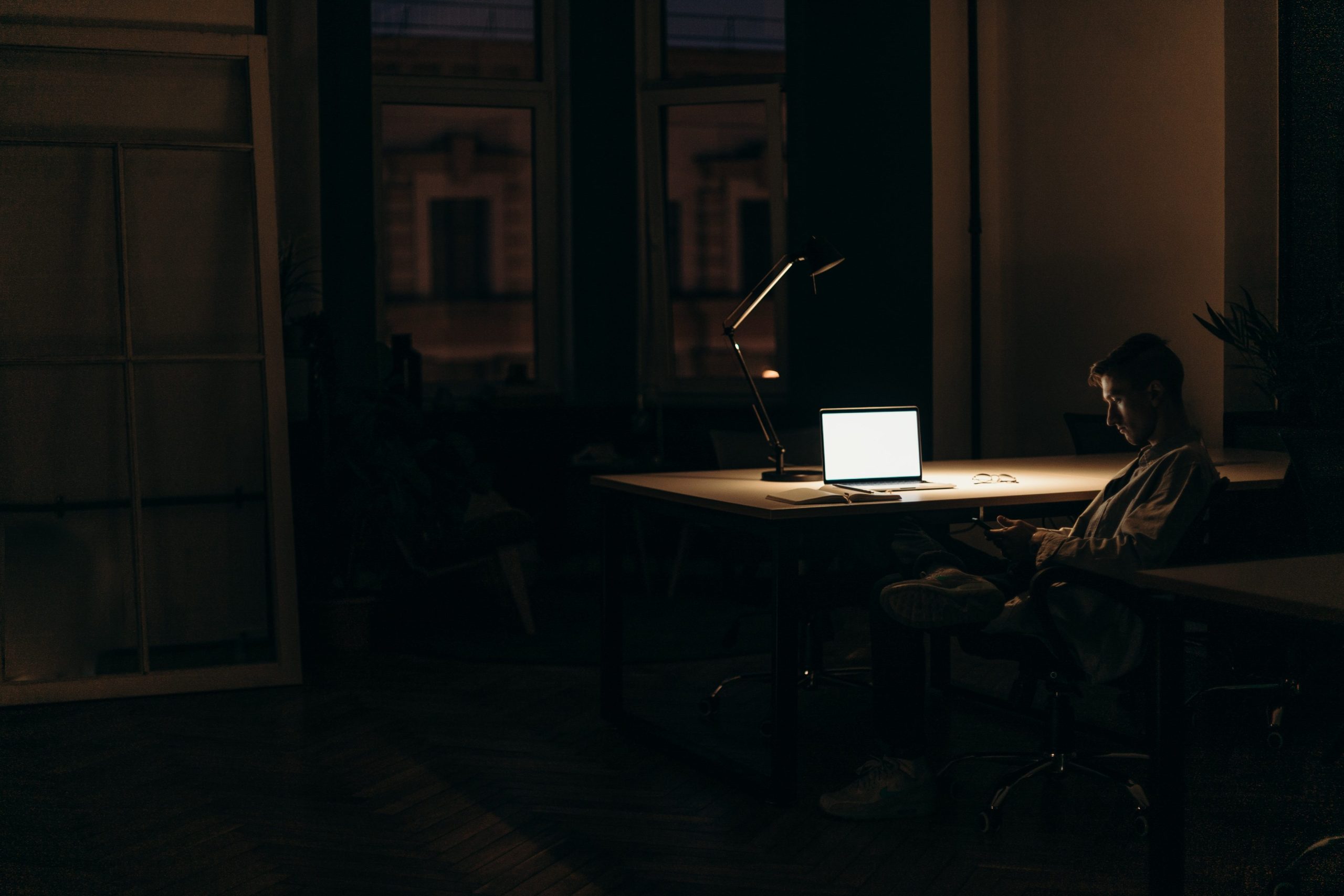 A man sits in front of a rectangular table in a dark room. He looking at his mobile phone. Over the table there is a computer, a notebook and a table lamp. The only light in the room comes from the lamp. The image is used to portrait inequalities in labour market exits, which are investigated in work package six.